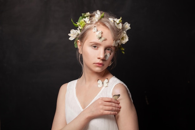 Märchen-Nymphe-Dame Schönes junges blondes Weibchen mit weißen Frühlingsblumen im Haar und Schmetterling auf ihrer gesunden Haut auf schwarzem Hintergrund