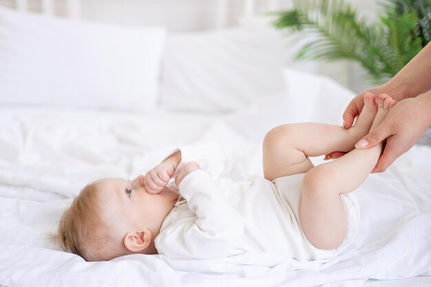mães mãos segurar as pernas bebê 6 meses de idade menino loiro em uma cama branca em um quarto brilhante depois de dormir de manhã em um bodysuit de algodão o conceito de bens infantis