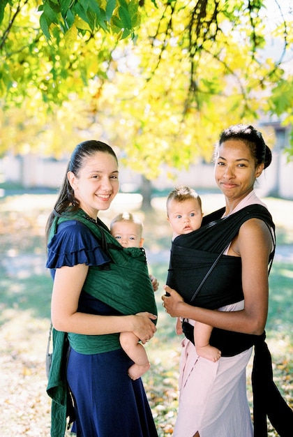 Mães sorridentes com bebês em slings sob uma árvore