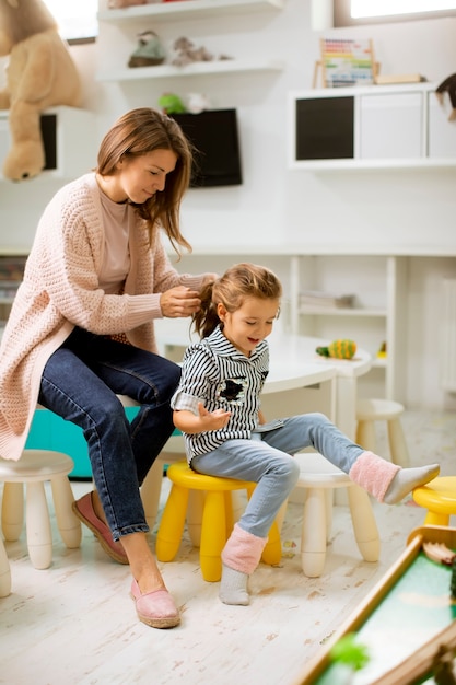 Maestra de guardería ayudando a la niña linda a arreglar el cabello en la sala de juegos del jardín de infantes