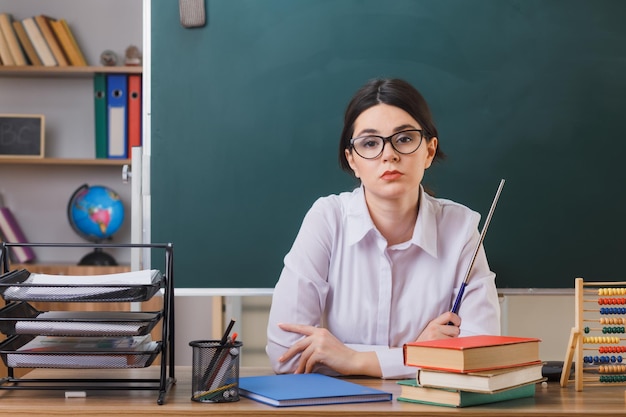 Una maestra joven y estricta con gafas sosteniendo un puntero sentada en el escritorio con herramientas escolares en el aula