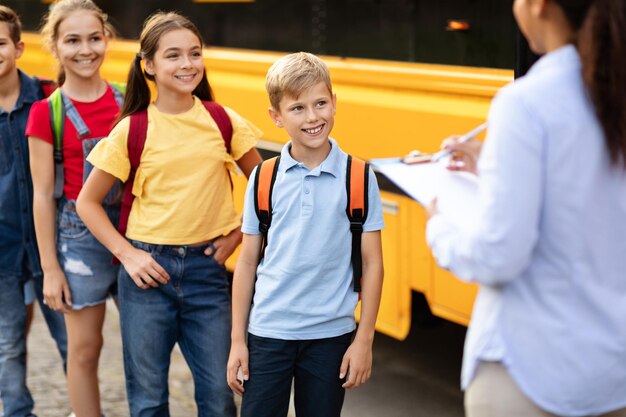 Foto maestra negra marcando nombres mientras los niños suben al autobús escolar