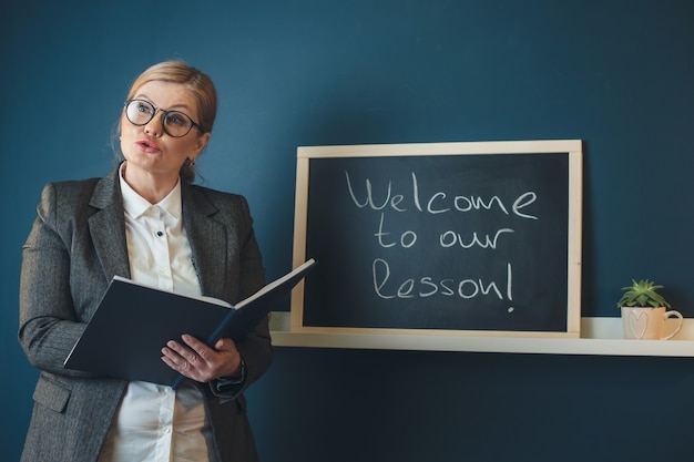 La maestra rubia senior está enseñando frente a la clase cerca de la pizarra sosteniendo un libro