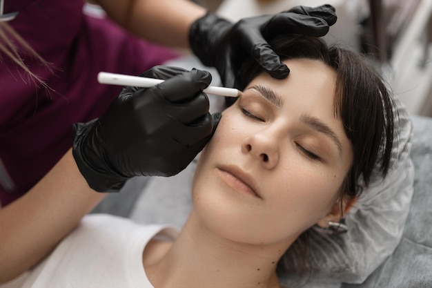 El maestro de cejas dibuja un contorno en las cejas del cliente con un lápiz blanco.