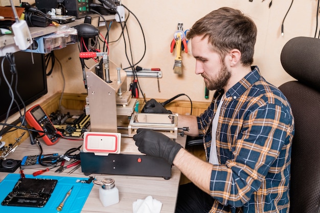 Maestro contemporáneo del servicio de reparación de dispositivos en guantes protectores con equipo especial mientras repara un teléfono inteligente