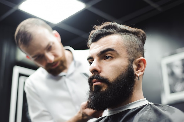 Maestro corta el pelo y la barba de los hombres en la barbería, el peluquero hace el peinado para un hombre joven.