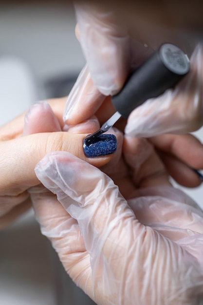 El maestro de la manicura sostiene el dedo del cliente y aplica un esmalte de gel azul en la uña.