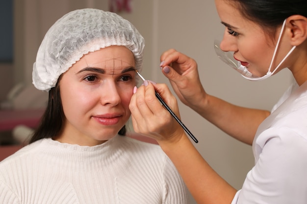 El maestro de maquillaje de cejas hace la preparación del diseño de la línea. Asistente es modelo de aprendizaje. Uniforme blanco, estéril. Arquitectura con herramientas.