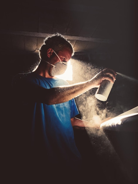 Maestro masculino en respirador rociando aerosol mientras muere tabla de surf en un taller de artesanía oscuro iluminado por la luz del sol que brilla a través del agujero