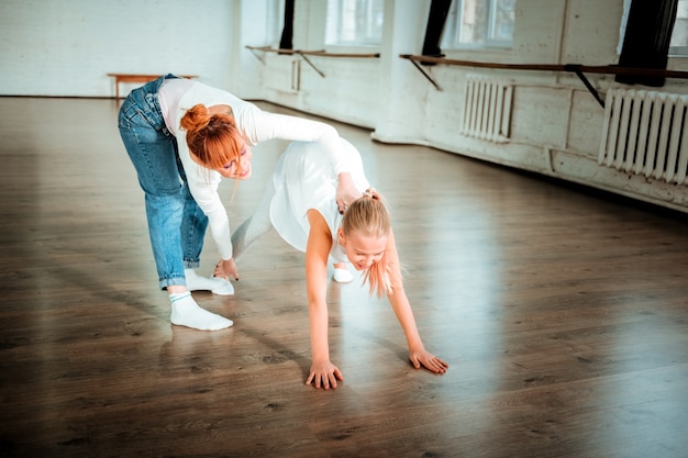 Maestro positivo. Profesora de ballet hermosa delgada con cabello rojo sonriendo amablemente mientras trabaja con su estudiante