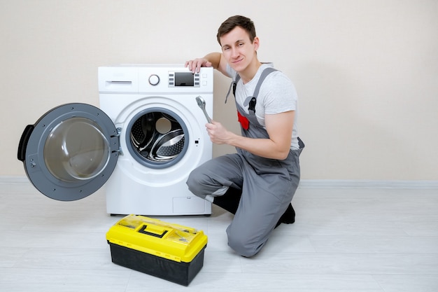 Maestro profesional con llave y caja de herramientas cerca de la lavadora moderna en el piso de madera cerca de la pared beige mirando a la cámara