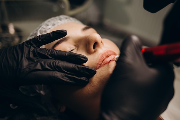 Maestro en salón de belleza pinta labios de chica modelo con máquina de maquillaje permanente Vista de cerca