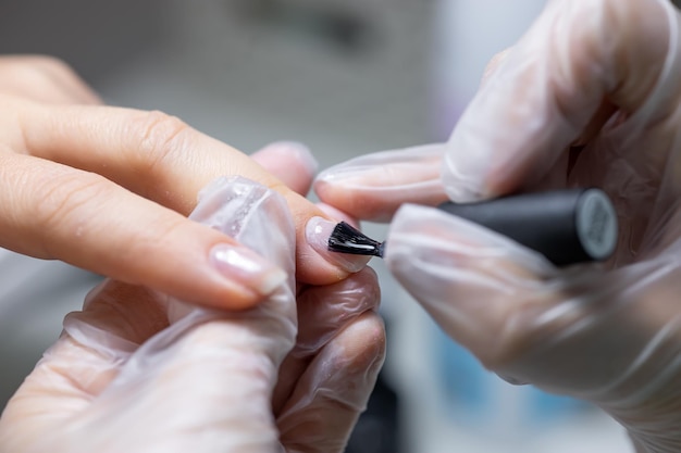 El maestro del servicio de uñas hace una manicura, sostiene su dedo y aplica un esmalte de gel transparente para proteger la uña.