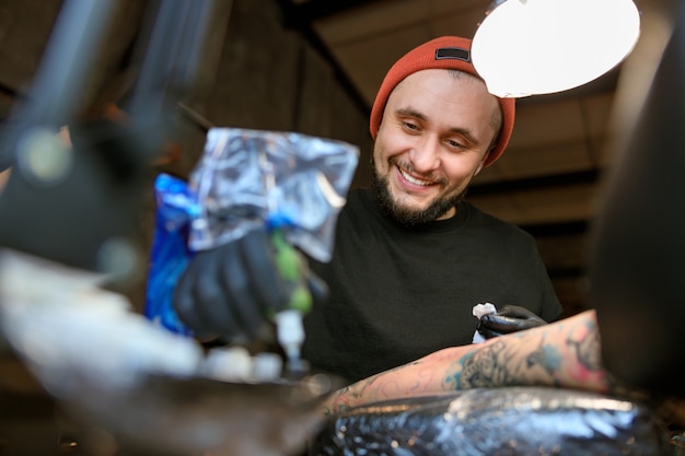 Maestro del tatuaje caucásico barbudo masculino profesional está trabajando en un acogedor estudio atmosférico