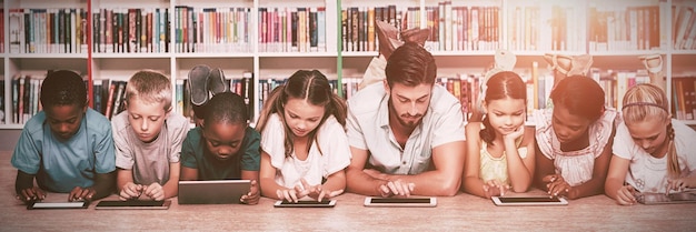 Maestros y niños tendidos en el piso con tableta digital en la biblioteca