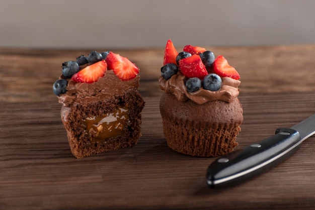 La magdalena de chocolate cortada está decorada con crema de mantequilla de chocolate y postre de bayas y un cuchillo en una tabla marrón de madera áspera