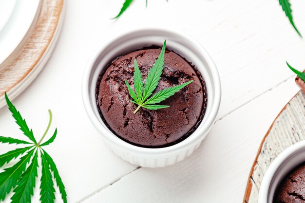 Magdalenas de chocolate con marihuana y hojas de cannabis, cbd de marihuana. Drogas de cáñamo de marihuana medicinal en postres de comida. Cocinar muffins de marihuana en una fuente para hornear con marihuana en la mesa de madera blanca Primer plano.