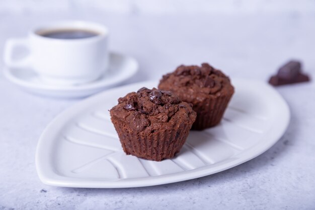 Magdalenas de chocolate en un plato blanco. Horneado casero. En el fondo hay una taza de café. Fondo blanco. Enfoque selectivo, de cerca.