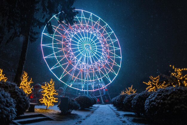 Magischer Winterpark mit Fahrgeschäften und erstaunlich leuchtendem Neon-Riesenrad bei Nacht mit Schnee