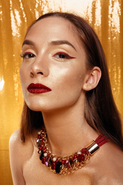 Magisches Mädchen-Porträt in Gold. Goldenes Make-up, Nahaufnahmeporträt in Studioaufnahme, Farbe. Beauty Model Girl mit perfektem hellem Make-up, roten Lippen, goldenem kastanienbraunem Schmuck. Sexy Lady Make-up Urlaubsparty.