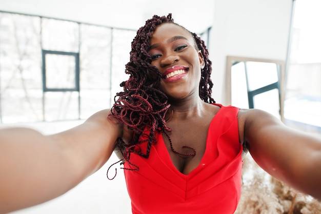 Magnífica joven africana con un lujoso vestido rojo sostiene la cámara y se hace selfie en un apartamento de lujo. Belleza, moda.