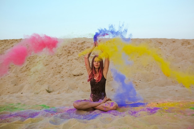 Foto magnífica modelo morena de cabelos compridos brincando com a pintura holi, sentada na areia