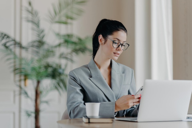 Magnífica mujer de negocios escribe mensajes en teléfonos inteligentes trabaja en nuevas poses de publicación frente a la computadora portátil involucrada en el proceso de trabajo se sienta en el escritorio bebe café aromático usa internet