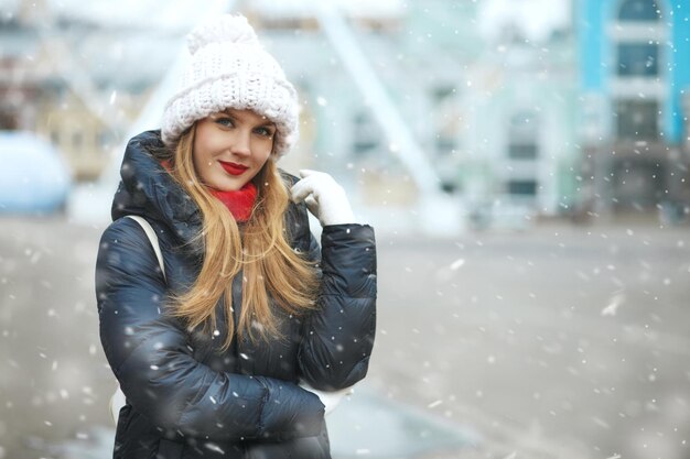 Magnífica mulher loira com boné de malha, caminhando na cidade durante a queda de neve. Espaço para texto