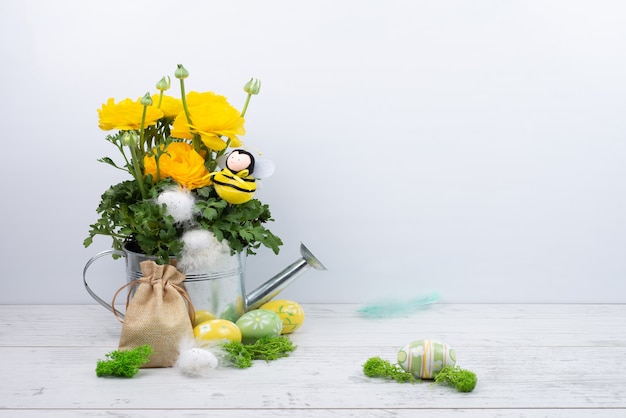 Magníficos ranúnculos amarillos del ranúnculo en una regadera de metal, abeja, huevos de Pascua con plumas, una bolsa de lona.