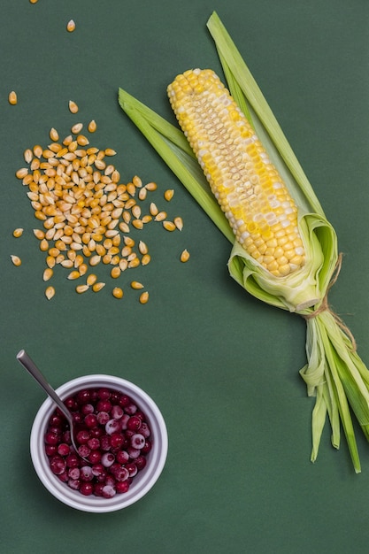 Foto maiskolben mit abgeschnittenen kernen. maiskörner auf dem tisch. rote beeren in weißer schüssel. grüner hintergrund. flach legen