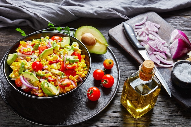 Maissalat mit Avocado, roten Zwiebelscheiben und Tomaten in einer schwarzen Schüssel auf einem dunklen Holztisch mit Zutaten auf dunklem altem Schneidebrett, mexikanische Küche, Landschaftsansicht von oben, Nahaufnahme