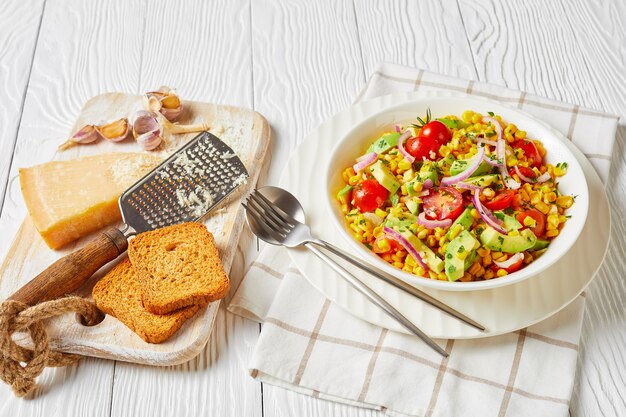 Maissalat mit Avocado, roten Zwiebelscheiben und Tomaten mit geriebenem Käse in einer weißen Schüssel auf einem Holztisch, Landschaftsansicht von oben