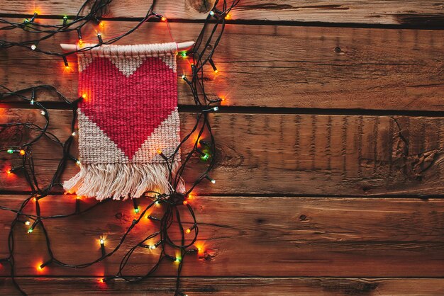 Makramee mit einem roten Herzen, umrahmt von einer Weihnachtsgirlande auf einem Holztisch. Ein Geschenk zum Valentinstag.