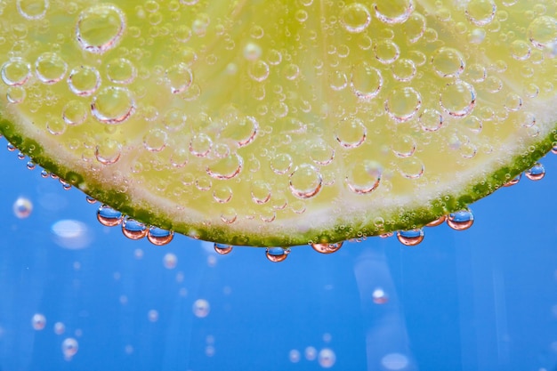 Makroaufnahme der gebogenen Kante einer durchsichtigen Limettenscheibe auf blauem Hintergrund mit sprudelnden Blasen