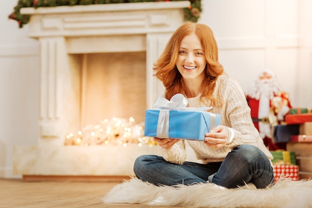 Mal posso esperar para abri-lo. Radiante senhora madura sentada no tapete fofo e sorrindo amplamente enquanto segura um presente e aproveita a manhã de Natal em casa.