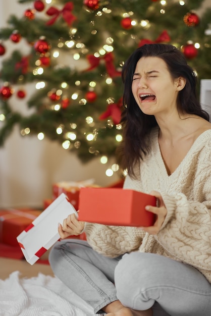 Mal regalo de Navidad mujer decepcionada e infeliz con caja de regalo abierta en el interior de casa