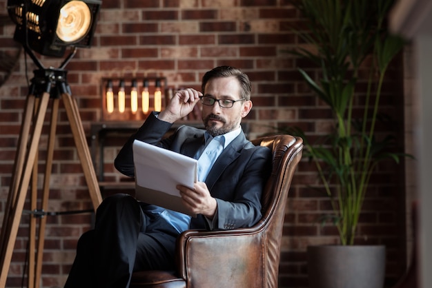 Mala vista. Agradable hombre adulto serio sentado en el sillón y sosteniendo sus gafas mientras lee sus notas