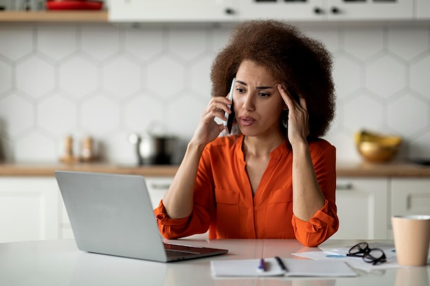 Las malas noticias estresaron a la mujer negra mirando la computadora portátil y hablando por teléfono celular