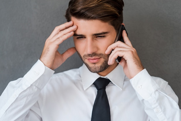 Malas noticias. Hombre joven frustrado en camisa y corbata hablando por teléfono móvil y tocando su cabeza mientras está de pie contra el fondo gris