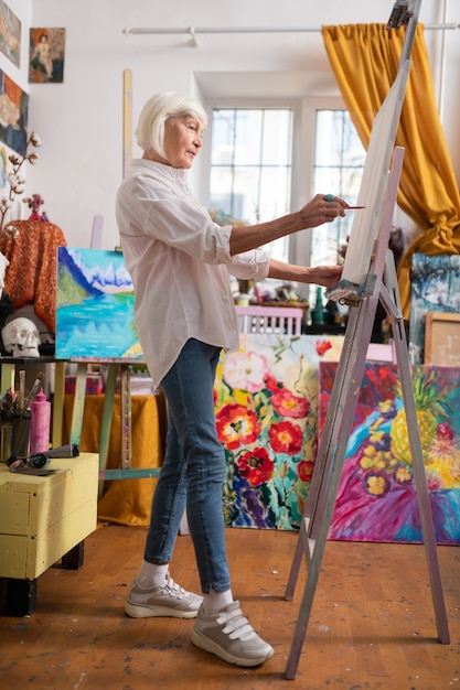 Malen in der Werkstatt. Alter Künstler, der Jeans und Turnschuhe trägt, die inspirierte Malerei in der Werkstatt fühlen