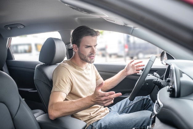 Malentendido. Grave joven adulto sentado al volante del coche gesticulando desconcertante durante el día