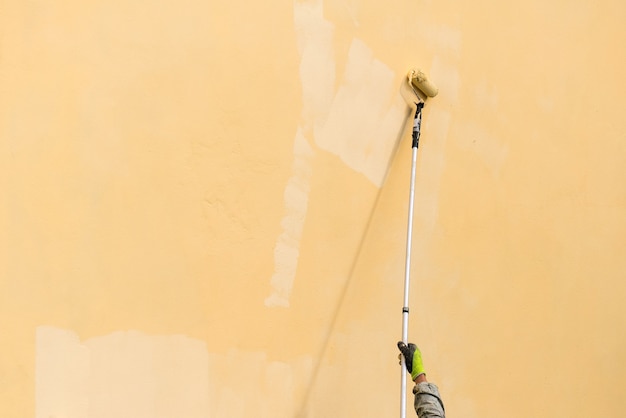 Maler malt Gebäudeaußenwand mit einer Rolle. Walze mit langem Stiel manuell Gebäude mit gelber Farbe streichen