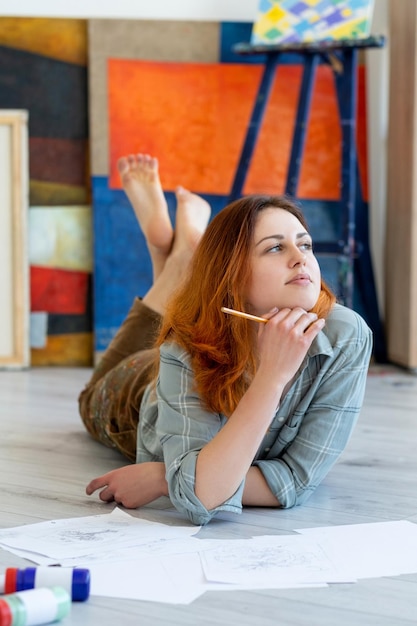 Malerei Kunst Künstler Lebensstil Talent Fantasie Inspirierte, nachdenkliche, entspannte Malerin, die barfuß mit Bleistiftskizzen im Lichtstudio mit orange-blauer abstrakter Kunstwerksammlung auf dem Boden liegt