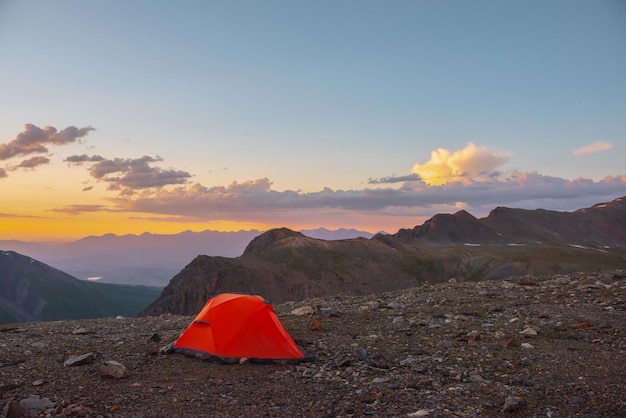 Malerische Alpenlandschaft mit Zelt in sehr großer Höhe mit Blick auf große Berge im orangefarbenen Morgenhimmel Lebendiges orangefarbenes Zelt mit herrlichem Blick auf die hohe Bergkette unter bewölktem Himmel in Sonnenuntergangsfarben