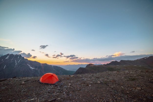 Malerische Alpenlandschaft mit Zelt in sehr großer Höhe mit Blick auf große Berge im orangefarbenen Morgenhimmel Lebendiges orangefarbenes Zelt mit herrlichem Blick auf die hohe Bergkette unter bewölktem Himmel in Sonnenuntergangsfarben