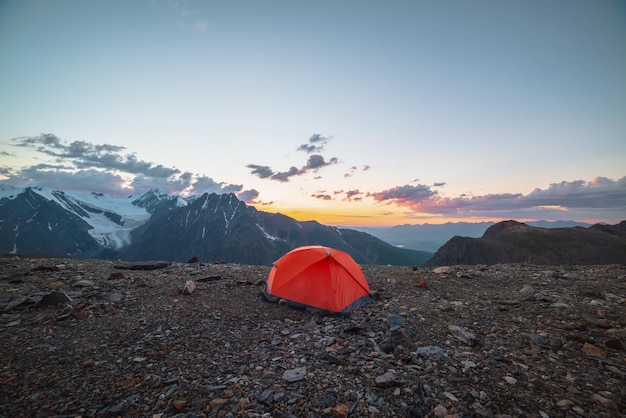 Malerische Alpenlandschaft mit Zelt in sehr großer Höhe mit Blick auf große Berge im orangefarbenen Morgenhimmel Lebendiges orangefarbenes Zelt mit herrlichem Blick auf die hohe Bergkette unter bewölktem Himmel in Sonnenuntergangsfarben