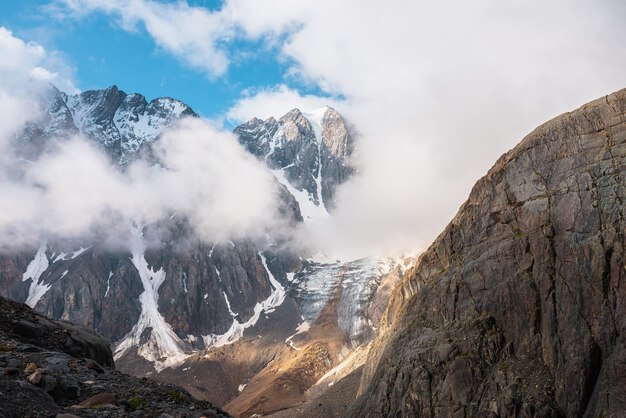 Malerische Berglandschaft mit schneebedeckter Bergspitze und Gletscher in dichten, niedrigen Wolken in Sonnenaufgangsfarben Farbenfroher Blick auf die Berge zum vertikalen Gletscher mit Eisfall im morgendlichen Sonnenlicht in dicken, niedrigen Wolken