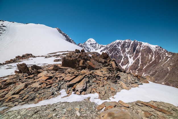 Malerische Berglandschaft mit sonnenbeschienenen Steinausreißern und hohen Schneebergen mit spitzem Gipfel unter blauem Himmel an sonnigen Tagen Ehrfürchtige alpine Landschaft mit scharfen Felsen und hohen schneebedeckten Gipfeln im Sonnenlicht