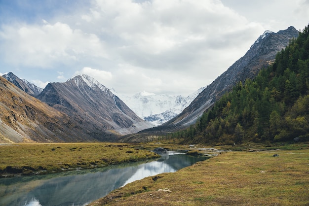 Malerische Herbstlandschaft mit Bergbach im Tal mit Blick auf großen Felsen mit Schnee oben und schöne hohe schneebedeckte Berge bei Sonnenschein. Stream mit klarem Wasser und tollen schneebedeckten Bergen.