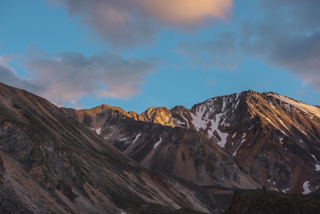 Malerische Landschaft mit einem hohen schneebedeckten Berg mit scharfem Felsgipfel im goldenen Sonnenlicht unter Wolken in Sonnenuntergangsfarbe bei wechselhaftem Wetter. Bunter Blick auf einen großen Berggipfel unter orangefarbenen Wolken am Himmel